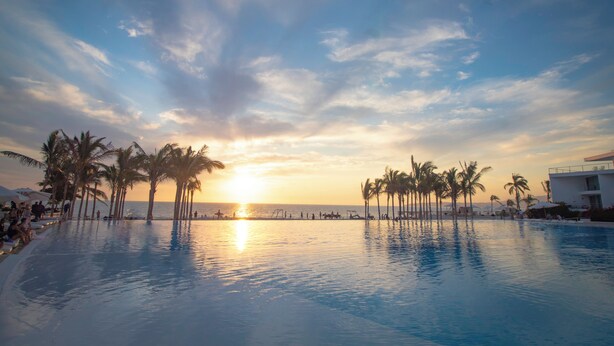 Vista a la piscina al atardecer, con palmeras y un edificio al fondo.