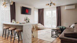 A modern living space with a kitchen island, bar stools, a sofa, and a TV displaying the Netflix logo. - DA-LI Residence - AC and Netflix (Brașov)