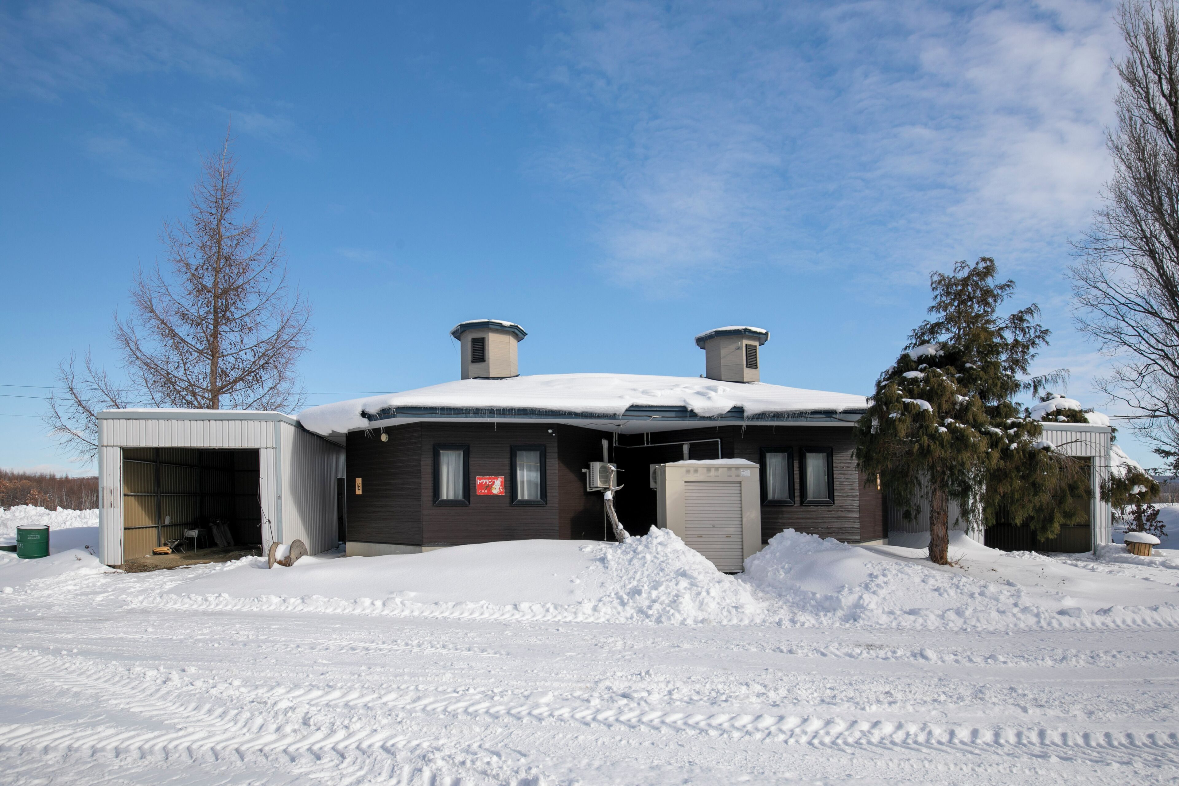 A two-story building with a snow-covered roof and a garage. There is a large pile of snow in front of the building, and a tree is visible to the right.