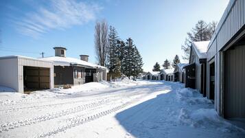 A snow-covered street with buildings on either side, including a garage and a building with a large tree in front.