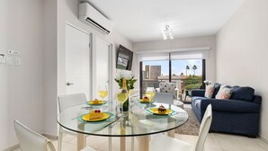 Ein modernes Wohnzimmer mit einem Glastisch, weißen Stühlen, einem blauen Sofa und Blick auf ein Gebäude sowie Palmen durch große Fenster.