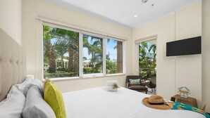 A hotel room with a large bed, a TV mounted on the wall, and a view of palm trees through the windows. - Amazing 3BR Eagle Beach Condo @LeVent  Pool  GYM (Noord)