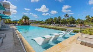 Une piscine entourée de palmiers, avec des chaises longues et des parasols, sous un ciel dégagé.