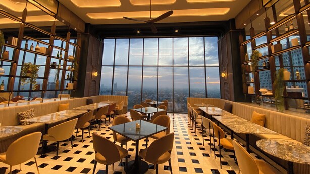 A modern restaurant with a high ceiling, large windows offering a city view, and a checkered floor pattern. - The Quarter Ratchayothin by UHG (Bangkok)