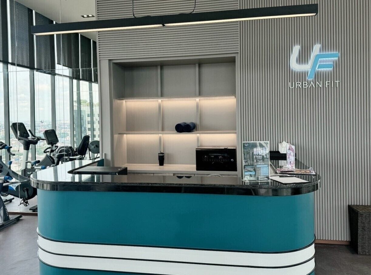 A modern reception desk with a teal and black color scheme, surrounded by exercise equipment and a large window offering a view of the city.