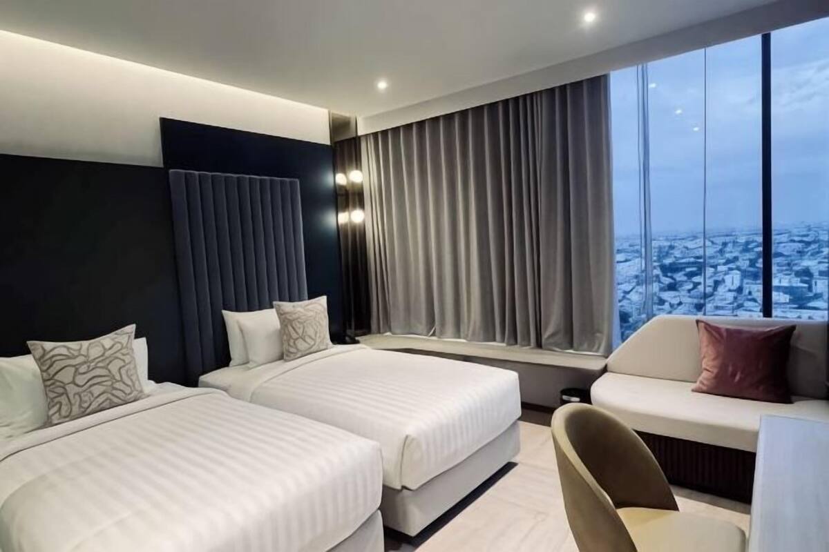 Una habitación de hotel moderna con dos camas, un sofá, un ventanal con cortinas y vistas a la ciudad.