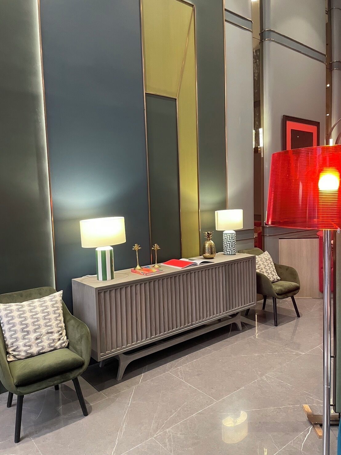 A modern hotel lobby with a central console table, two armchairs, a large mirror, and a red lamp.