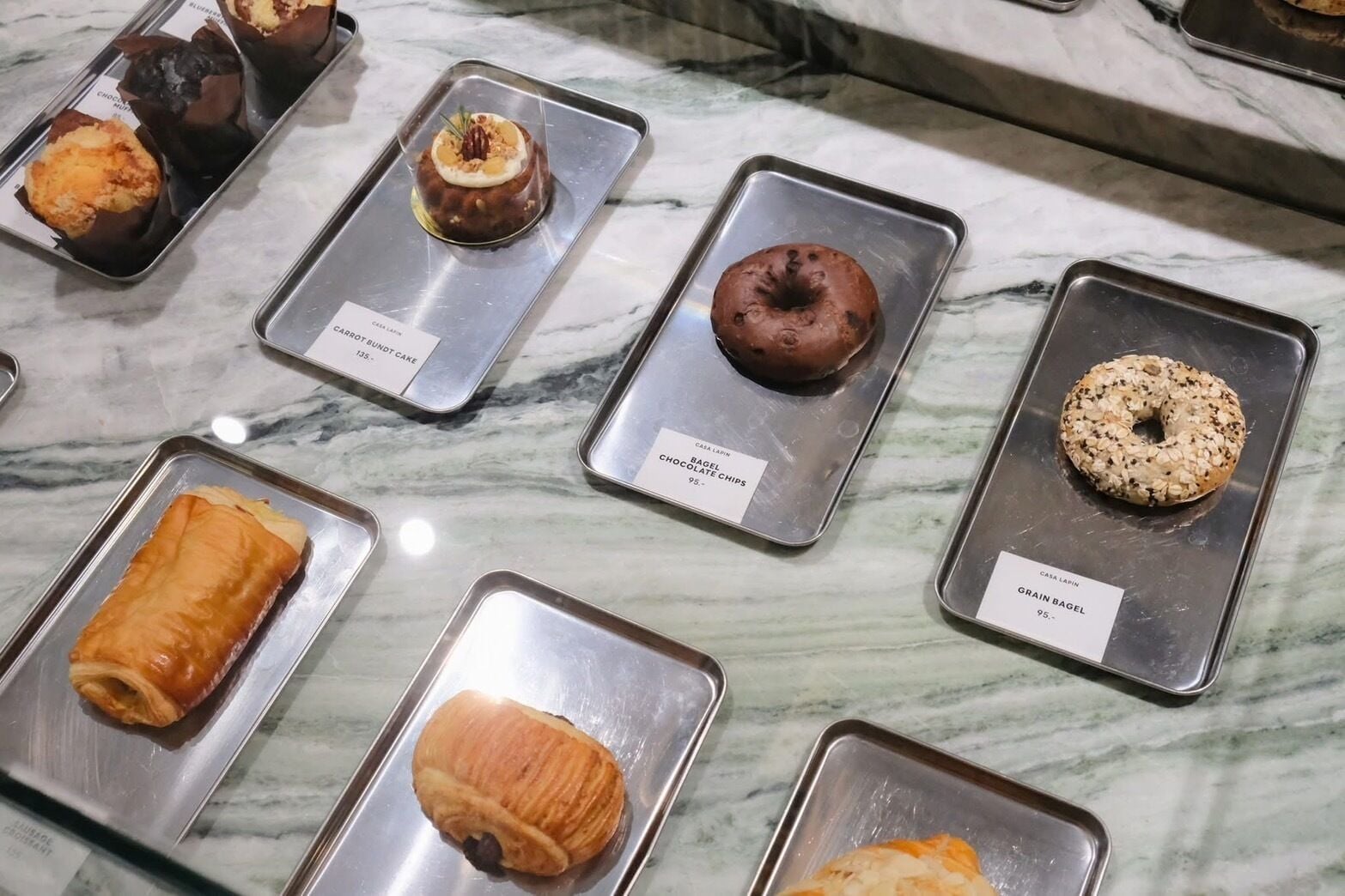 A selection of baked goods on trays with labels indicating their names and prices.