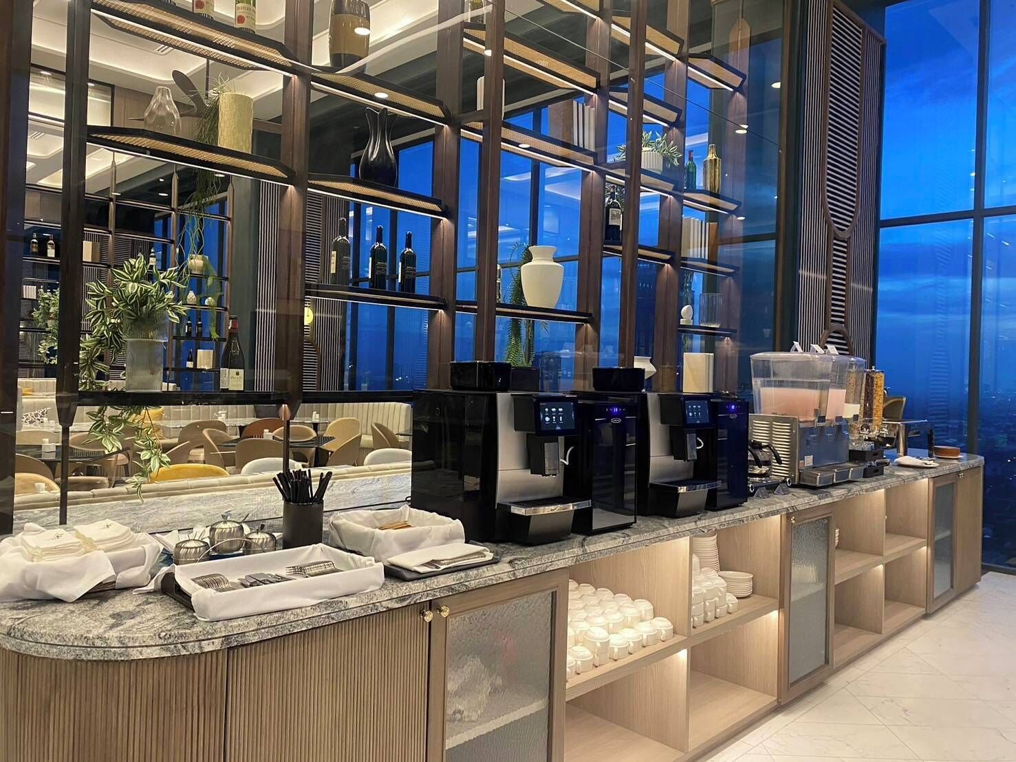 A modern bar area with a glass partition, coffee machines, and neatly arranged dining ware.