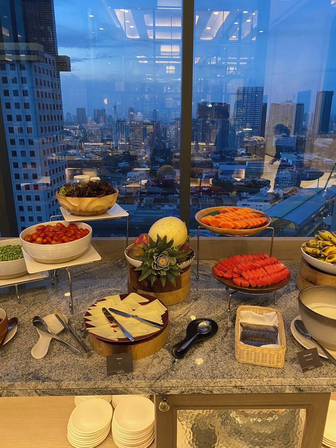 Een buffetopstelling met diverse gerechten en bestek, een stadszicht door een groot raam, en een modern interieur.