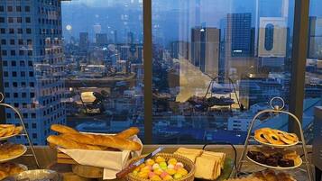 A breakfast buffet with various pastries and bread, set against a cityscape view through large windows.