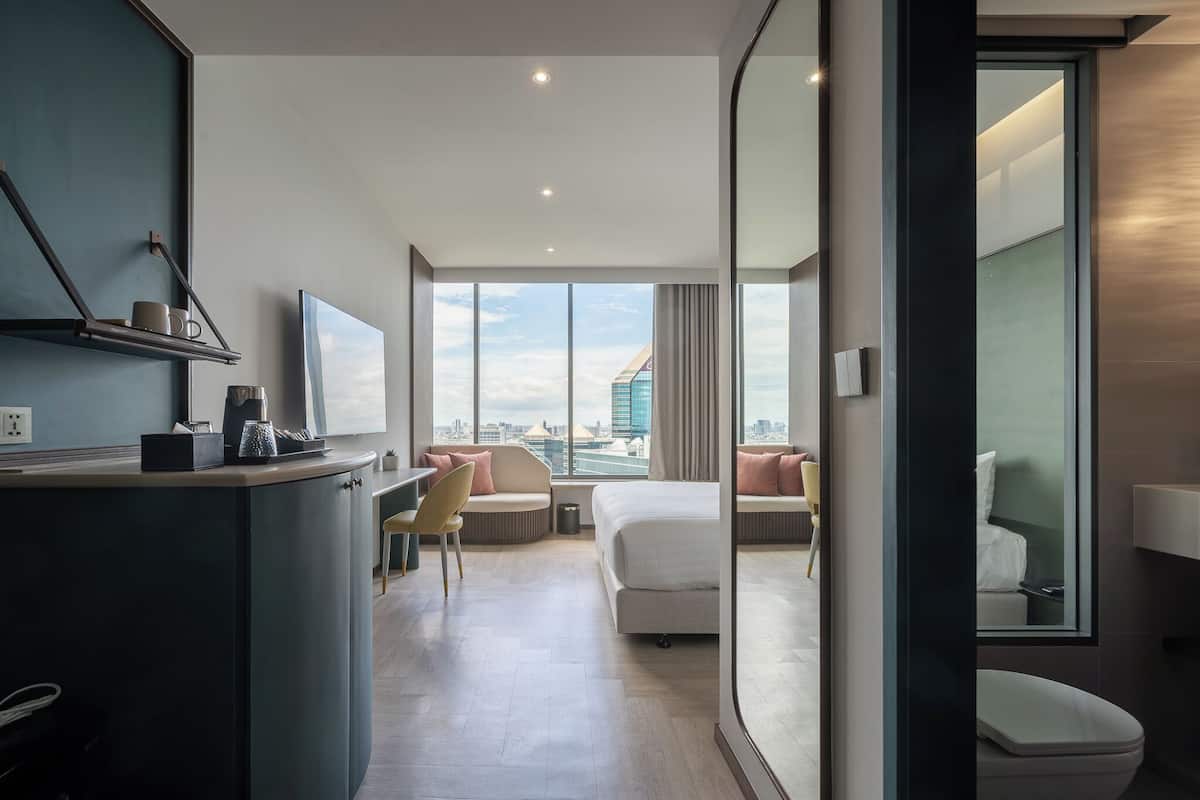 A modern hotel room with a desk, a bed, a sofa, and a large window offering a city view.