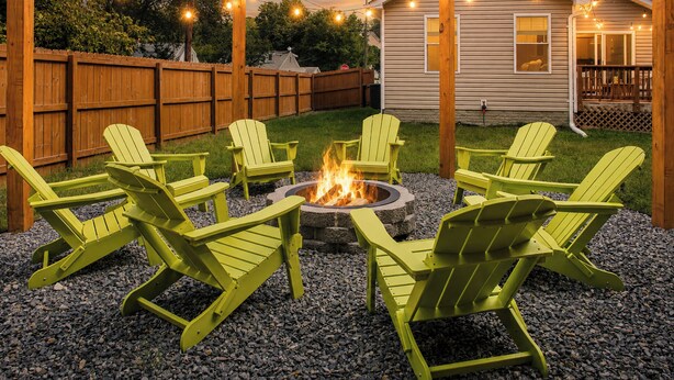 Un patio trasero con un brasero rodeado de sillas Adirondack verdes, una valla de madera y una casa con luces colgantes.