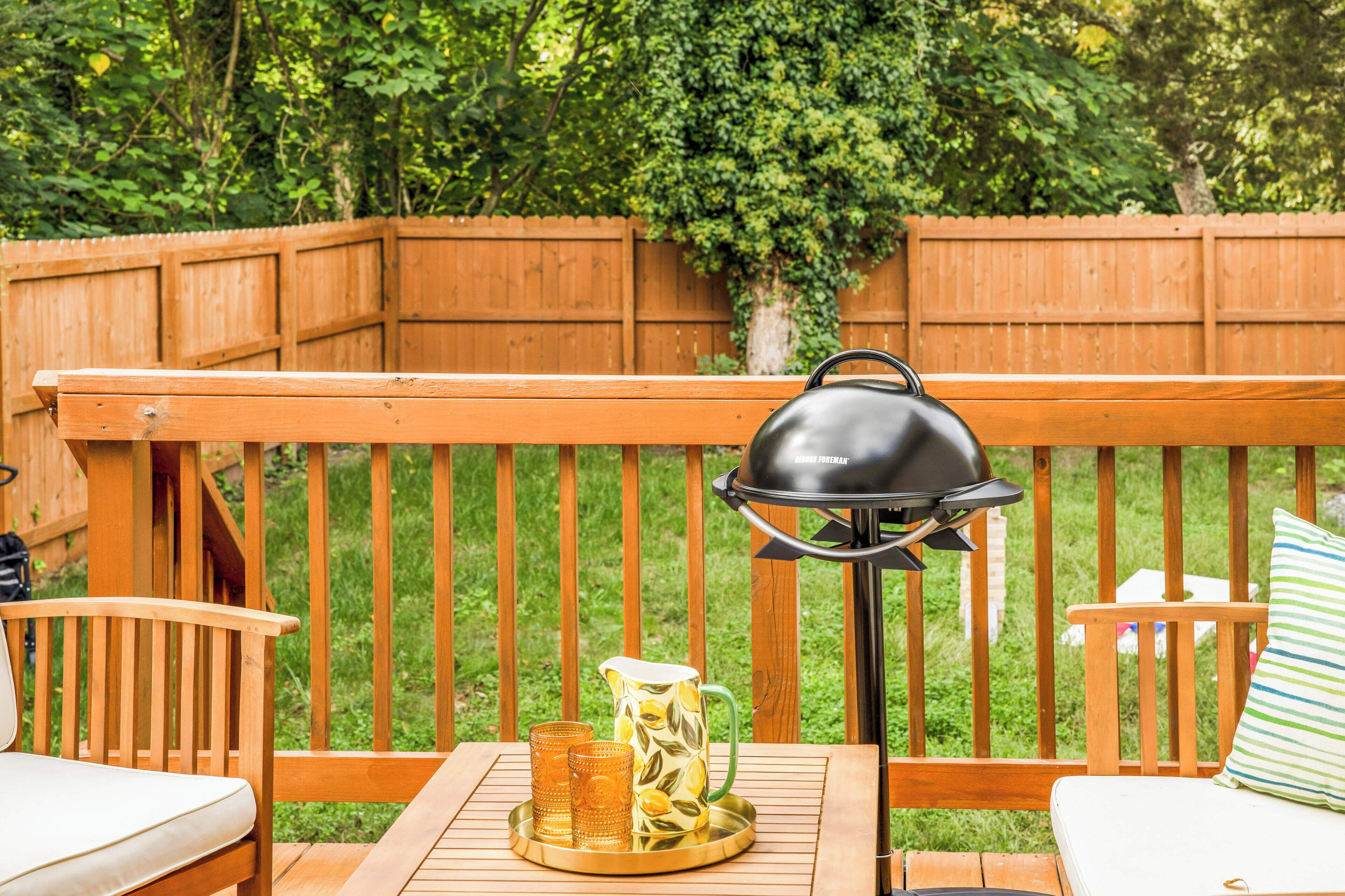 A wooden deck with a grill, a table with a pitcher and glasses, and a cushioned chair.