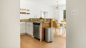 Una cocina moderna con armarios blancos, encimera de granito y lavavajillas. Hay un comedor con mesa blanca y sillas amarillas.