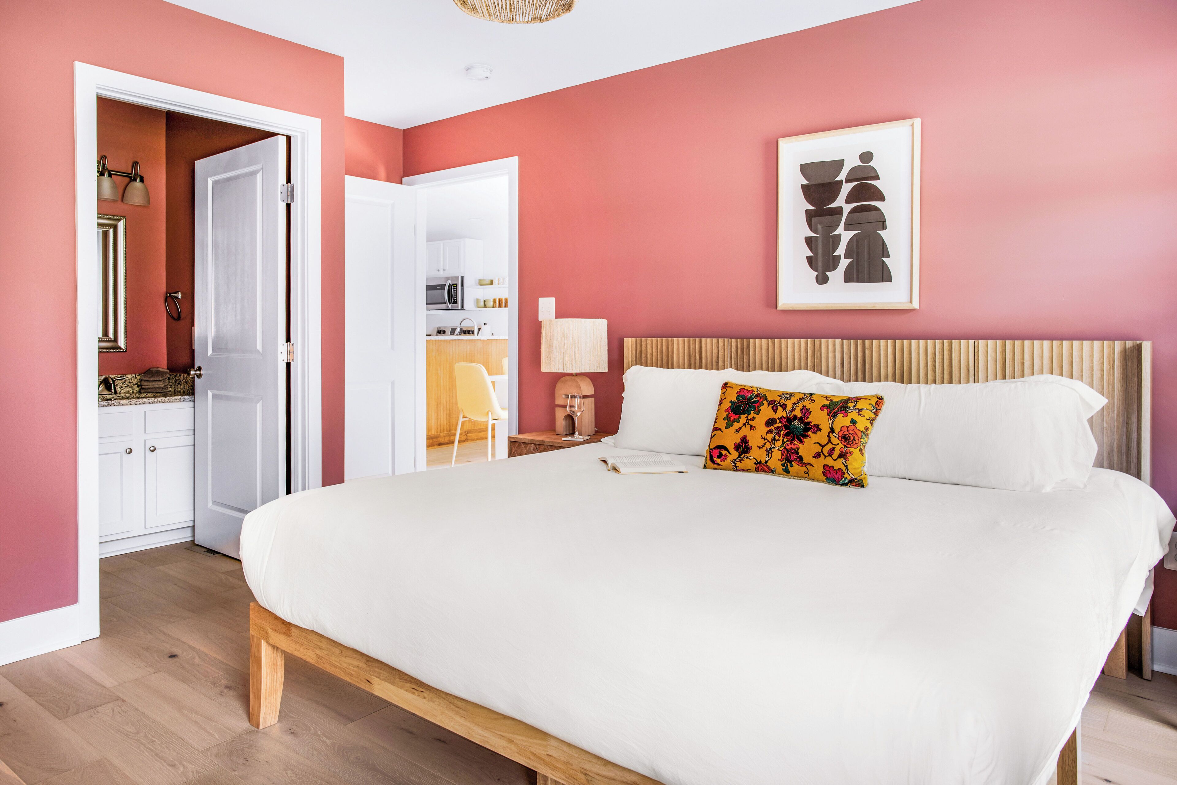 Une chambre avec un grand lit, une table de chevet avec une lampe, une porte donnant sur une salle de bain et une cuisine visible à travers une porte ouverte.