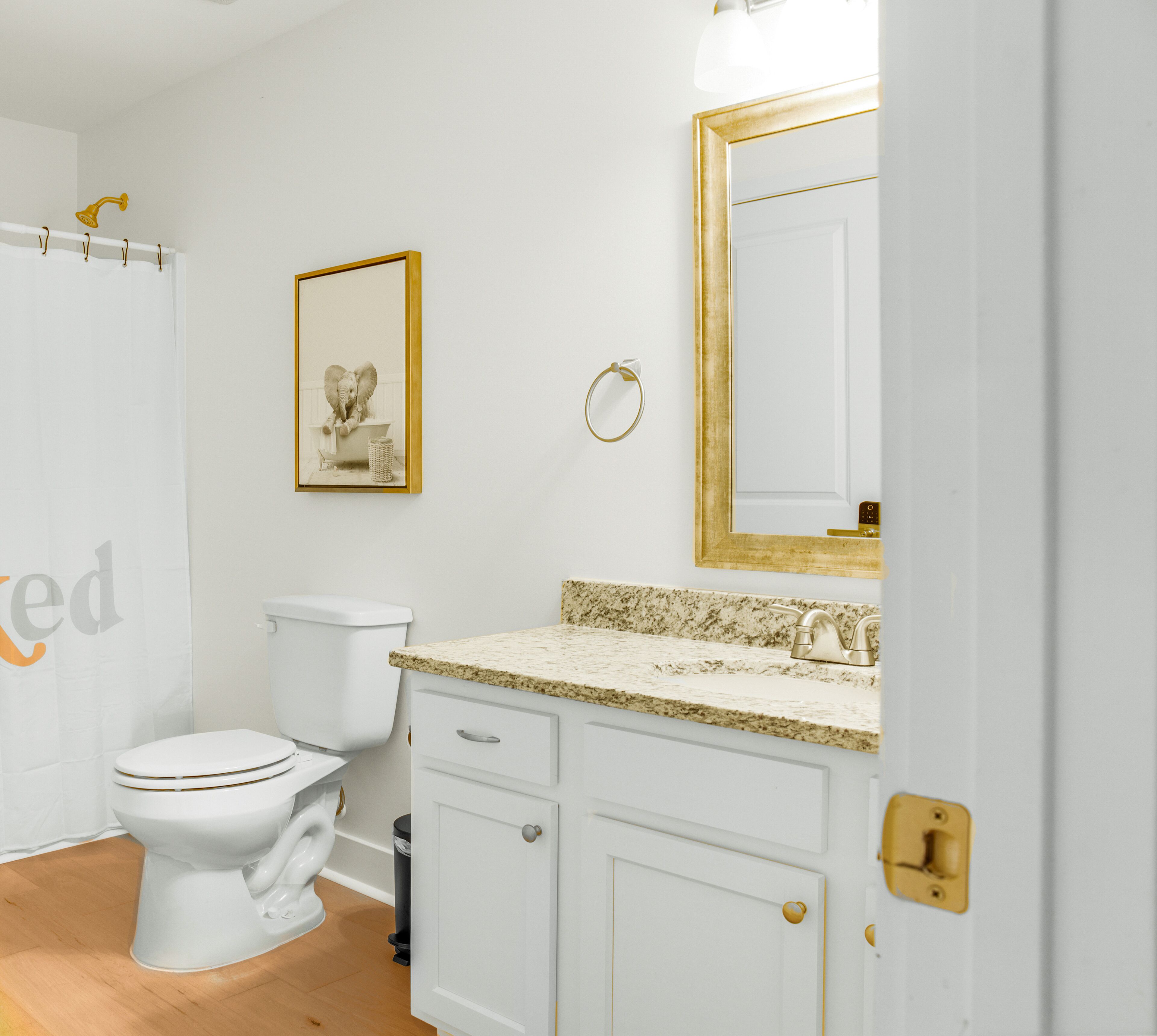 A bathroom with a toilet, a sink with a granite countertop, a mirror with a gold frame, and a shower curtain.