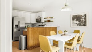 Una cocina moderna con armarios blancos, un refrigerador de acero inoxidable, un microondas e isla de madera. Hay una mesa de comedor con sillas amarillas y una lámpara colgante blanca.