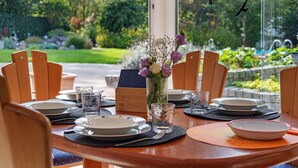 Mesa de comedor preparada con platos, vasos y un florero, rodeada de sillas, con vista a un jardín y un edificio al fondo.