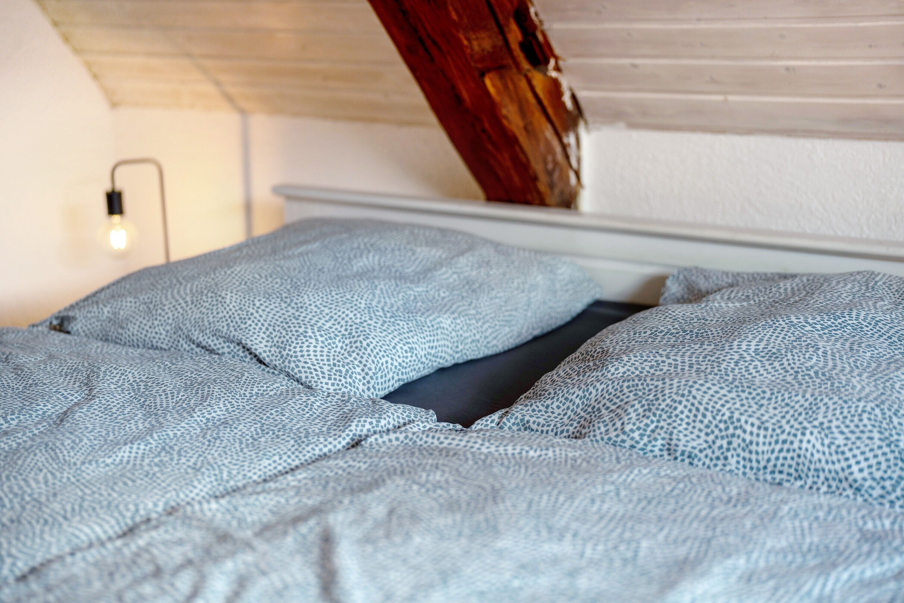 A bed with a patterned blanket, a wooden beam, and a wall-mounted light fixture.