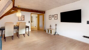Un espace repas avec une table et des chaises, un plafond avec des poutres apparentes, un téléviseur fixé au mur et une porte donnant sur une autre pièce.