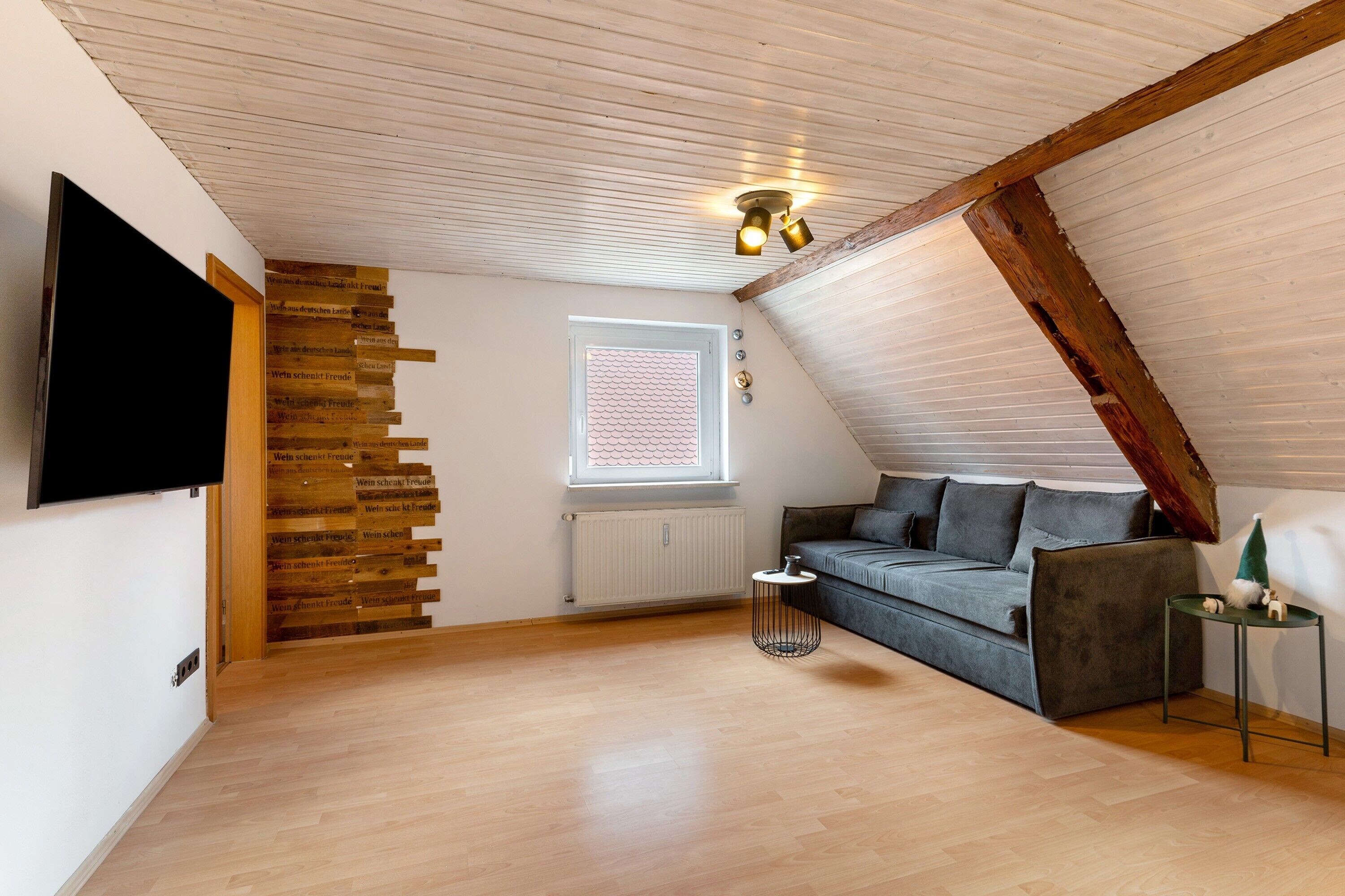 Una habitación estilo loft con techo inclinado, televisor empotrado, sofá gris, una mesita redonda y ventana con vistas.