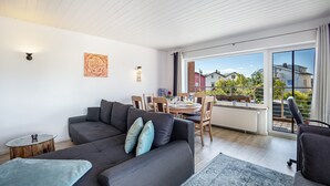 Een moderne woonkamer met een bank, een eettafel met stoelen, een klein bijzettafeltje en een groot raam met uitzicht op huizen.