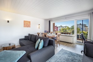 Ein modernes Wohnzimmer mit einer Couch, einem Esstisch und Stühlen, einem kleinen Beistelltisch und einem großen Fenster mit Blick auf Häuser.
