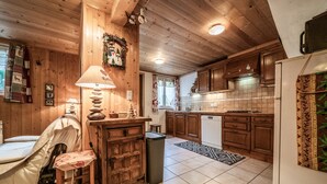 Un interior de madera con una cama, una silla, una lámpara, un cubo de basura, una zona de cocina con armarios, una estufa y un refrigerador.