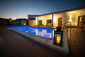 A swimming pool with lounge chairs and a table, surrounded by a building with a stone wall and a lantern.