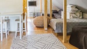 Una habitación moderna y luminosa, con suelo de madera, un escritorio blanco con dos sillas, un sofá, una cama y un televisor montado sobre una pared de ladrillo blanco.