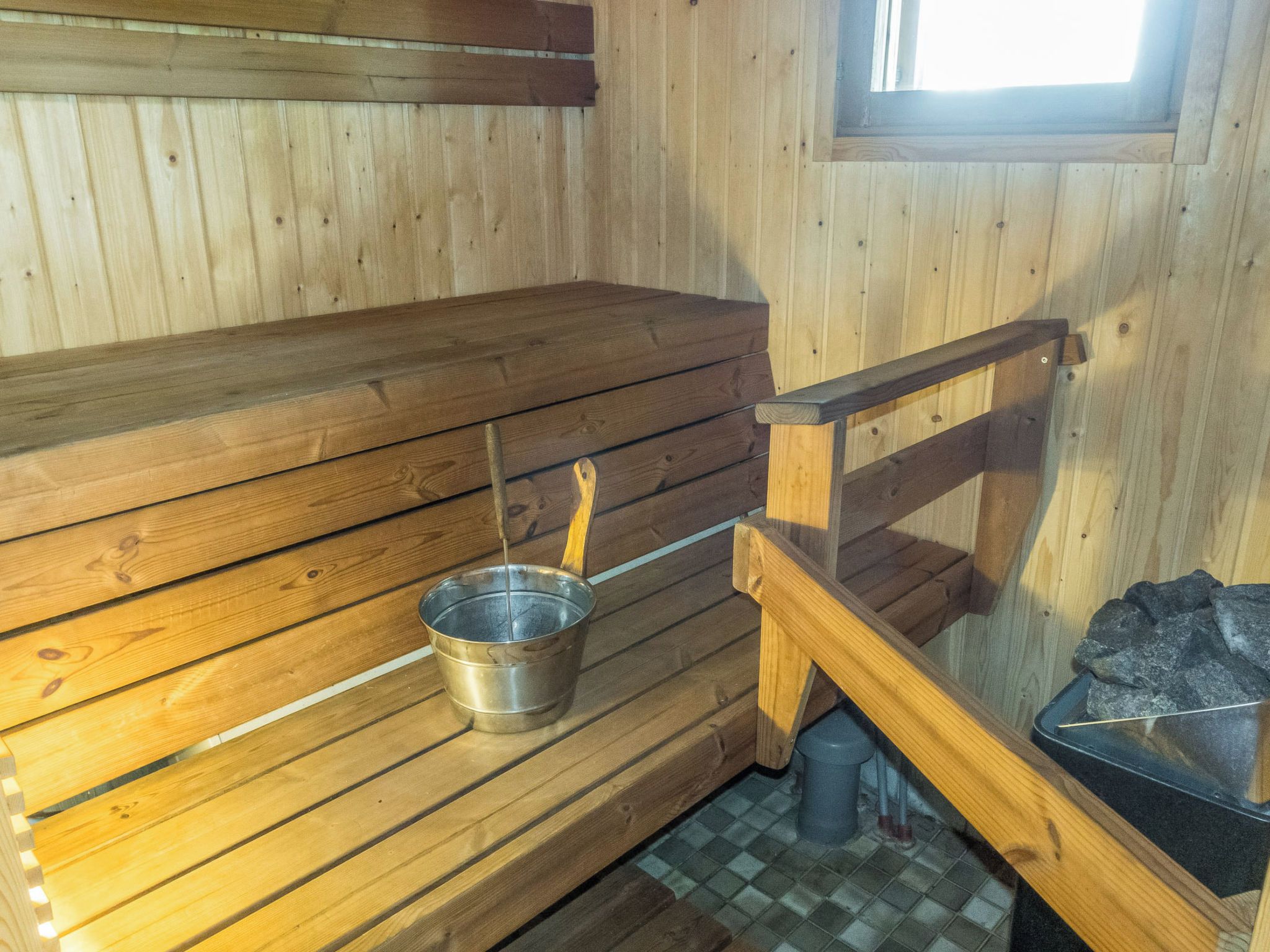 Una sauna con bancos de madera, un cubo metálico y un recipiente de piedra.