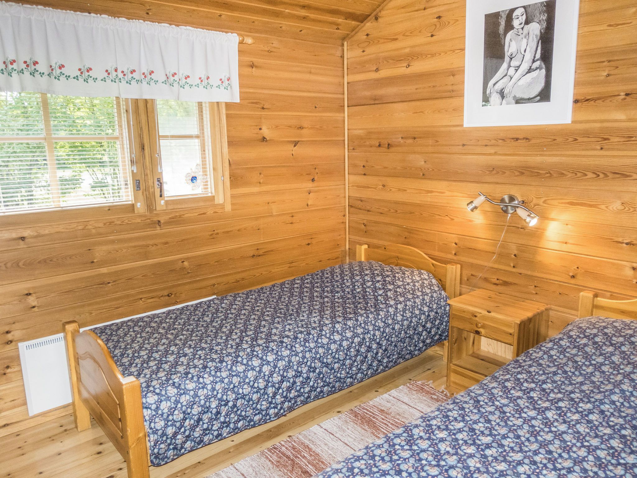 A small wooden room with two single beds, a window with blinds, a wall-mounted light, and a framed picture.