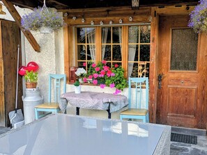 Een houten huisje met een glazen tafel en stoelen, een deur en bloemenmanden.