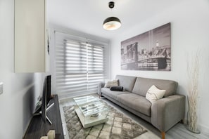 Un salon moderne avec un canapé, une table basse en verre, un téléviseur et un tableau représentant un paysage urbain.