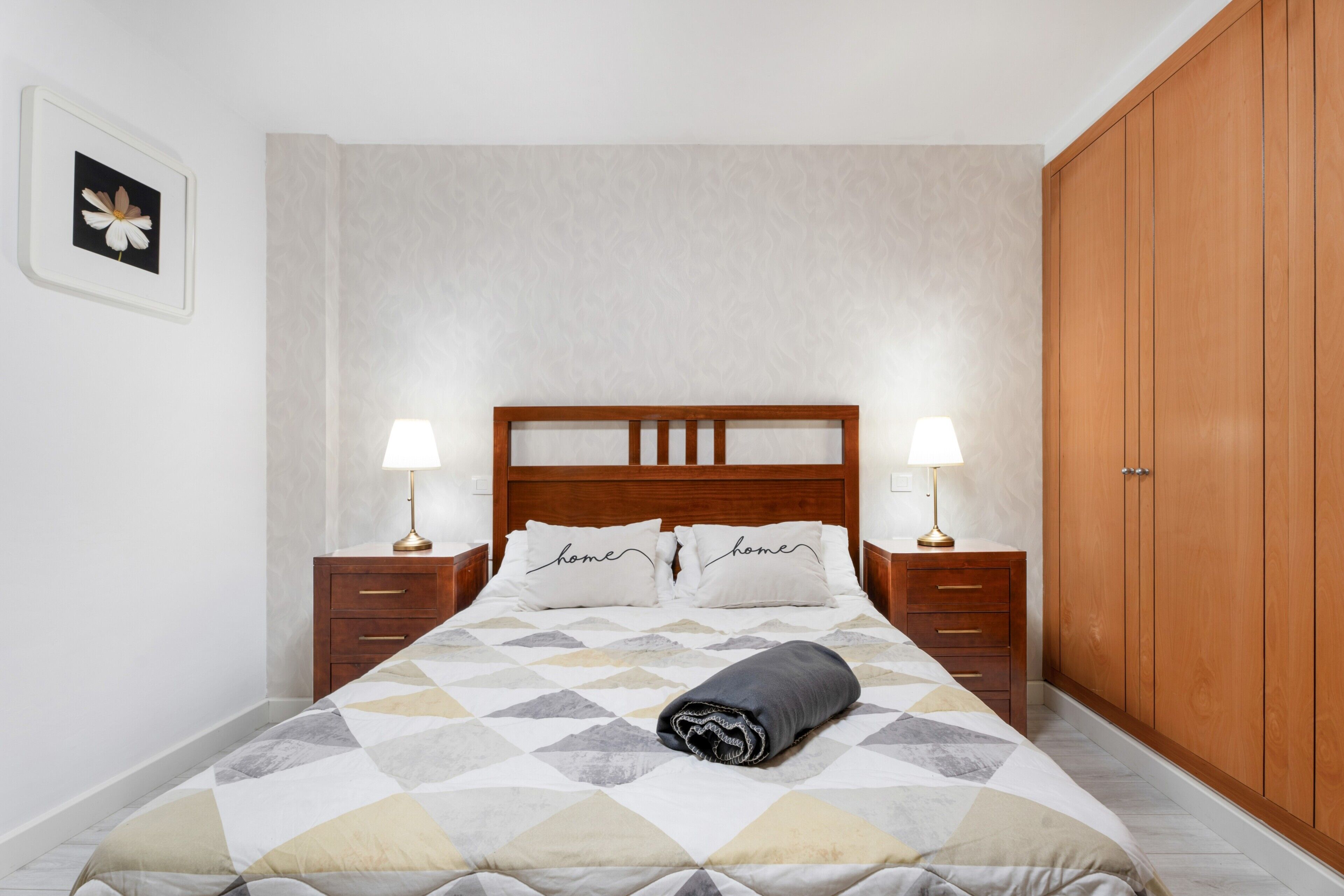 Un lit bien fait, avec une couvre-lit à motifs géométriques, deux lampes de chevet, une tête de lit en bois et une armoire en bois.