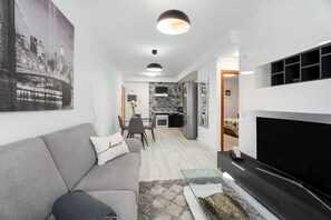 Una sala moderna con un sofá, una mesa de centro de vidrio, un televisor de pantalla plana y una cocina al fondo.