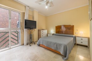 A bedroom with a bed, a television on a stand, a nightstand, a lamp, and a window with a view of a brick building.