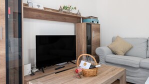 Una sala de estar con televisor de pantalla plana, una mesa de centro de madera, una cesta de mimbre con copas de vino y un sofá de color gris claro.