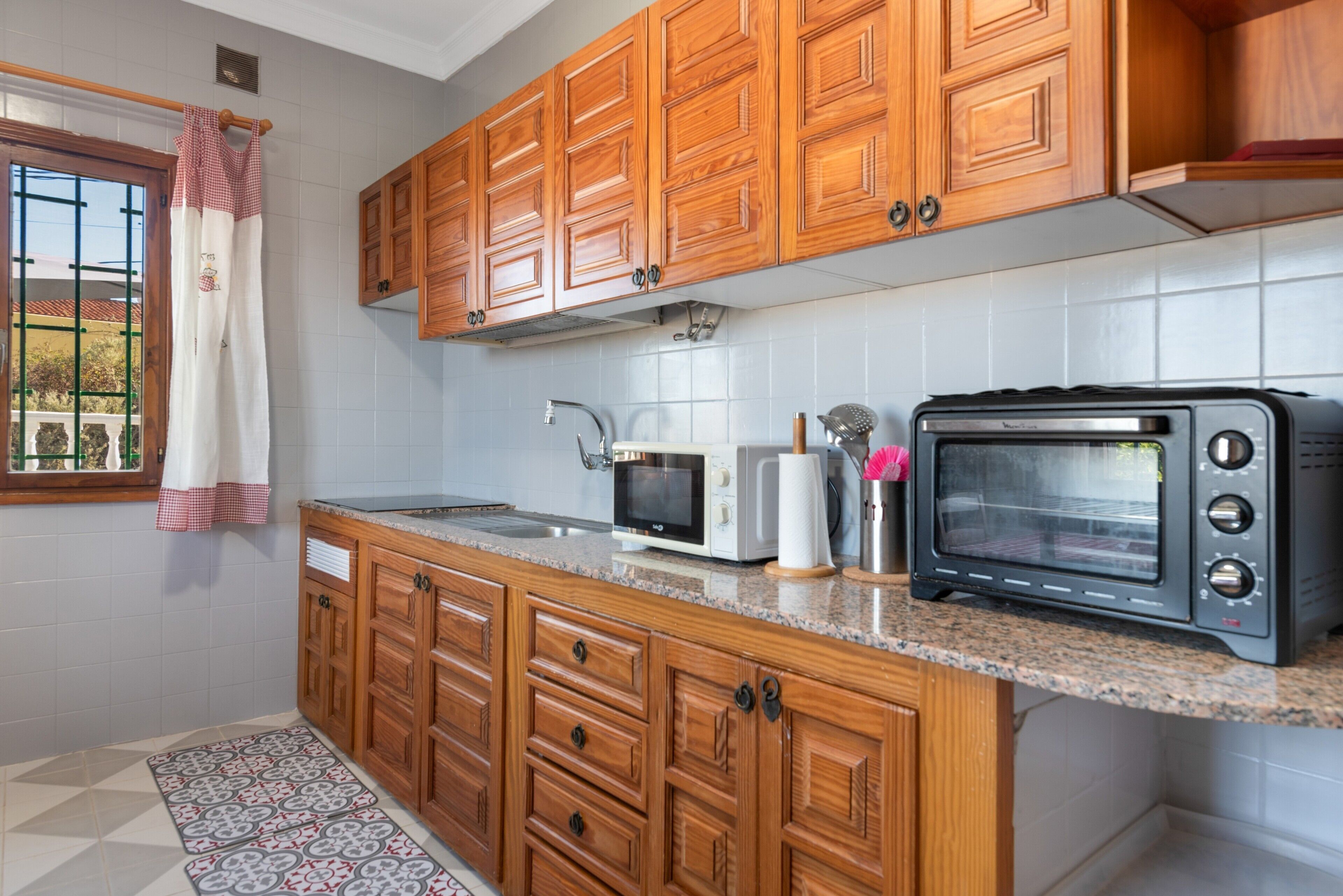 A kitchen with wooden cabinets, a microwave, a toaster oven, and a patterned rug.