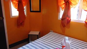 Quarto de hotel com cama listrada, cortinas laranja amarradas e uma pequena mesa, além de um quadro na parede.