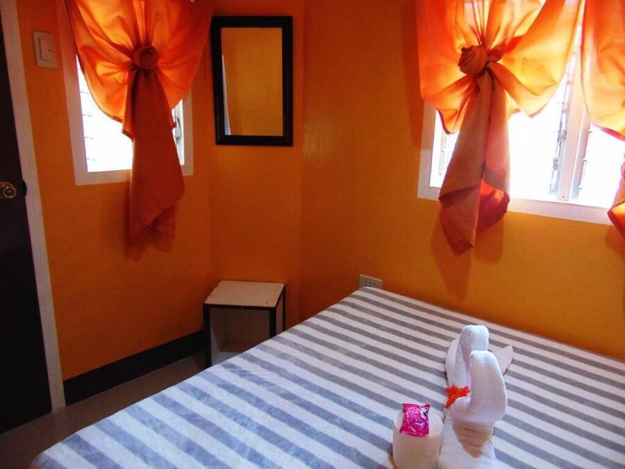 A hotel room with a striped bed, orange curtains tied back, a small table, and a framed picture on the wall.