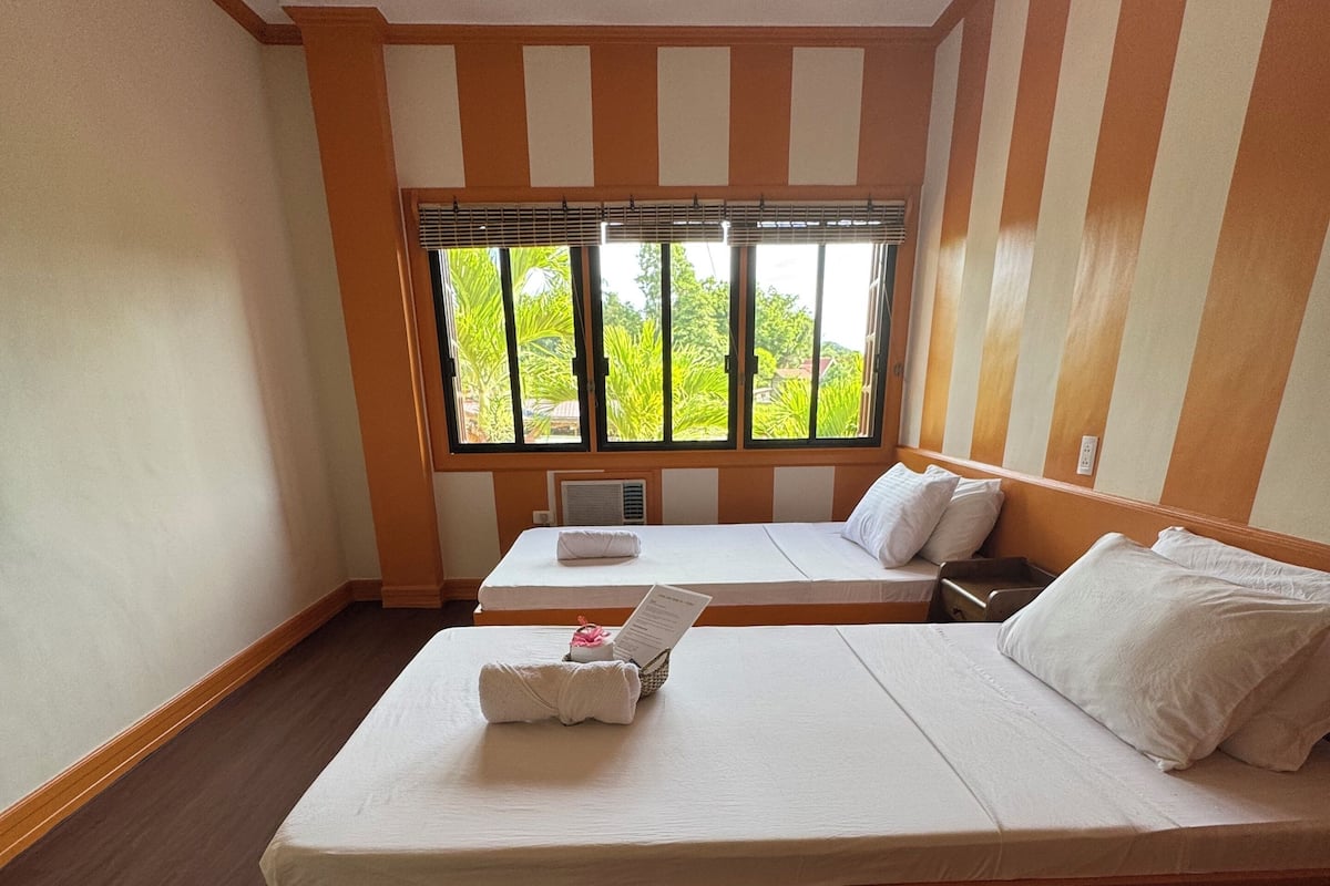 A hotel room with two beds, a window with blinds, a small table, and a wall with striped panels.