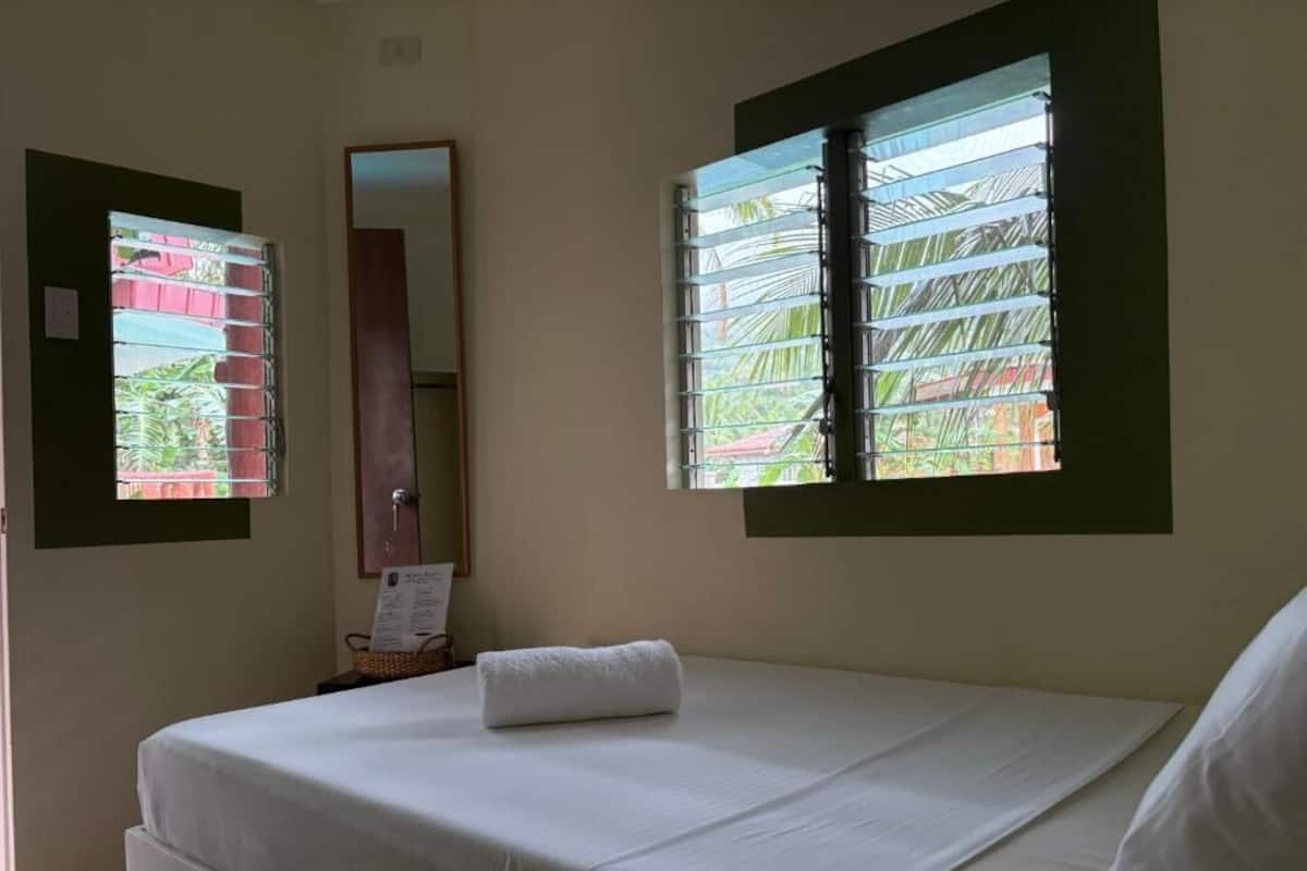 A room with a bed, a window with blinds, a mirror, and a wall-mounted light fixture.