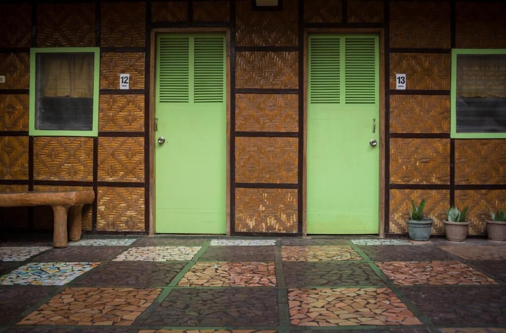 Fachada de edificio con dos puertas verdes, numeradas 12 y 13, y un banco a la izquierda.