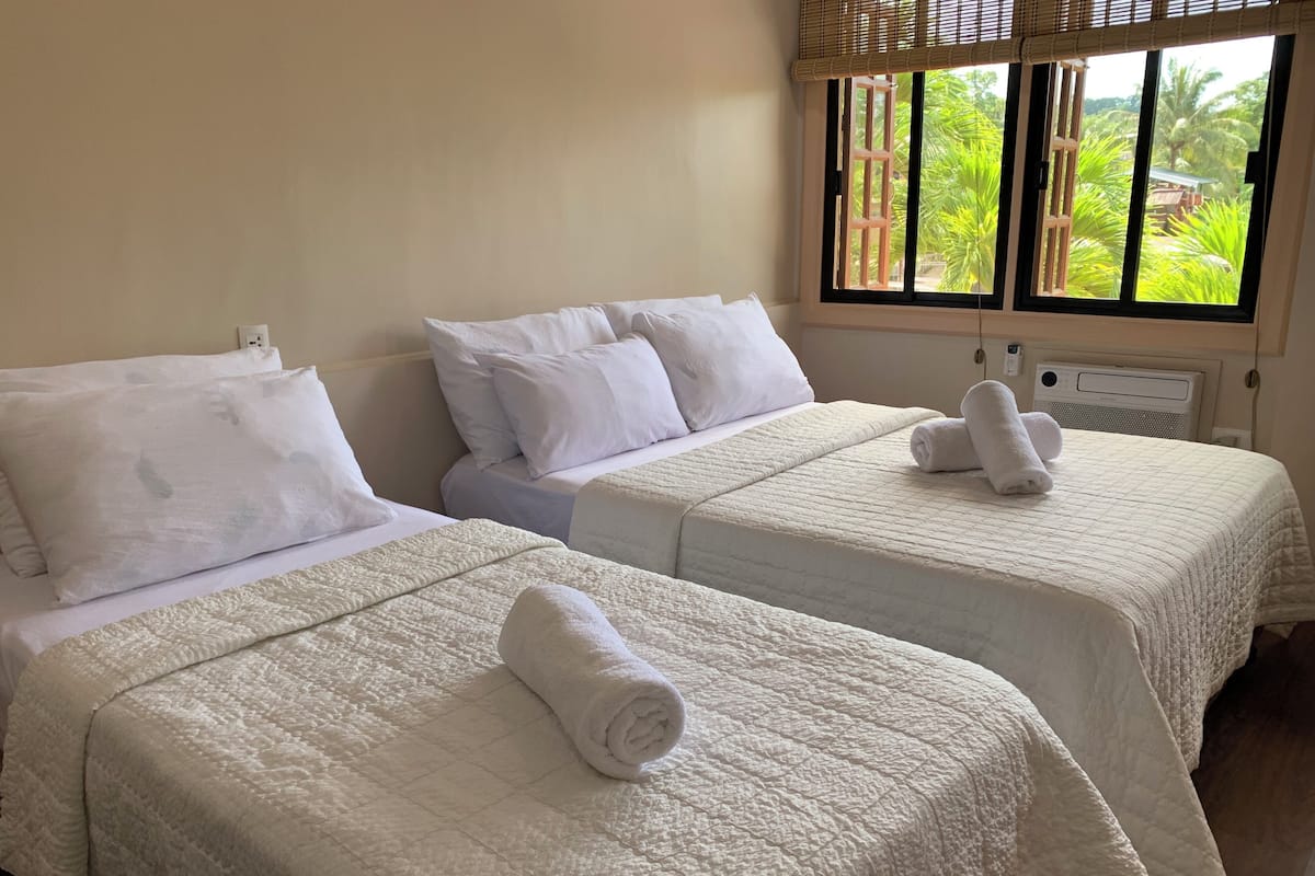 Two beds with white bedding, a window with a view of greenery, and a wall socket.
