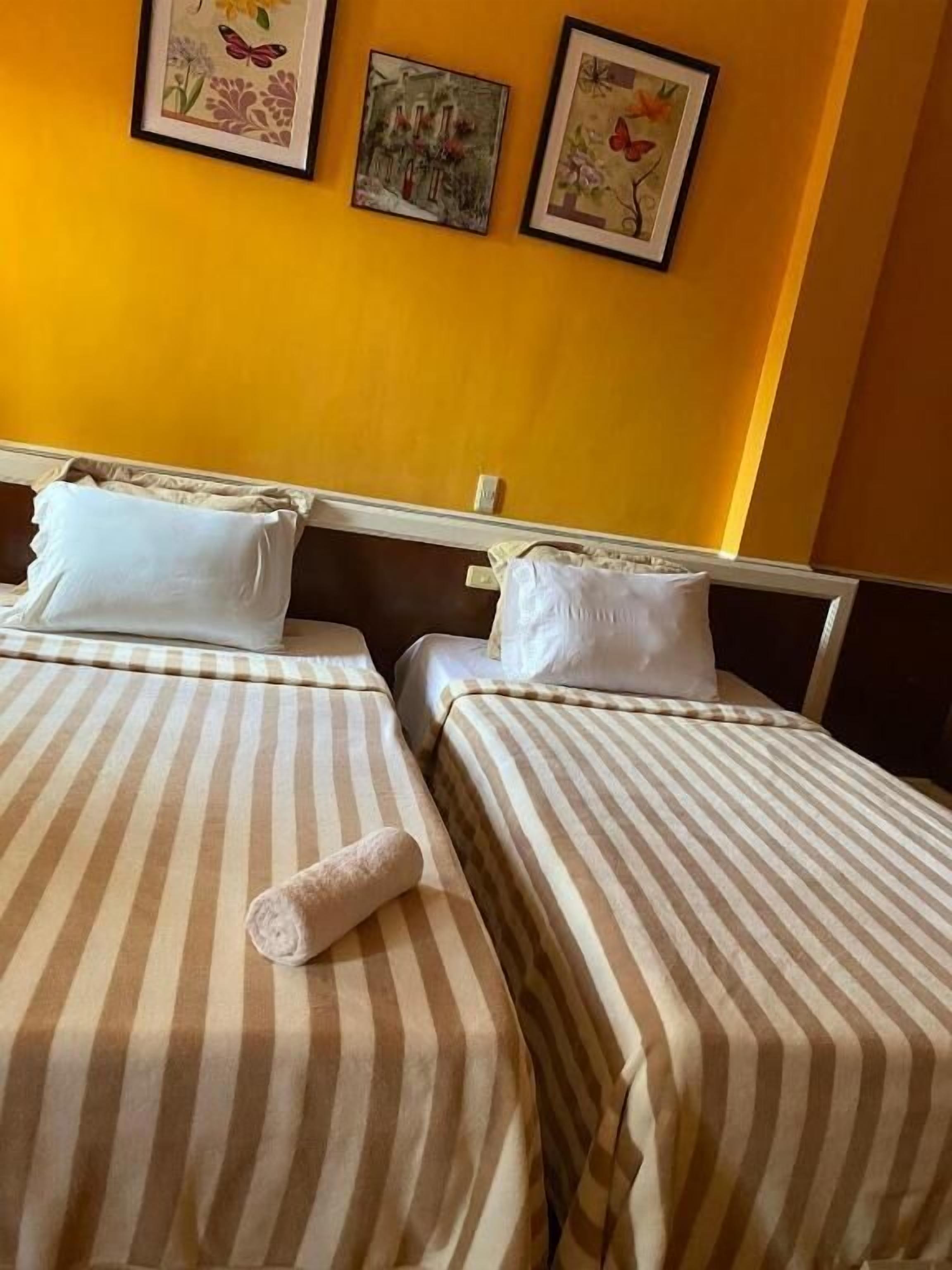 A hotel room with two beds, each with striped bedding and white pillows. There are three framed artworks on the wall above the beds.