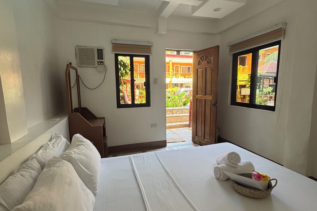 A hotel room with a bed, pillows, a bedside table, a lamp, and two windows.