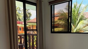 Une chambre d’hôtel avec un lit, des rideaux, une fenêtre donnant sur des immeubles et des palmiers, et un balcon.