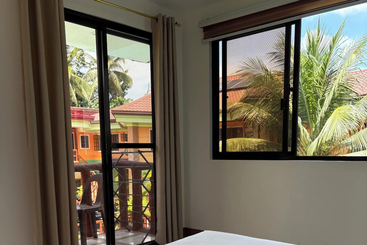 A hotel room with a bed, curtains, a window with a view of buildings and palm trees, and a balcony.